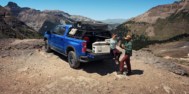 A women is seating on Silverado which is parked on top of hill and a men stands and enjoy a snacks